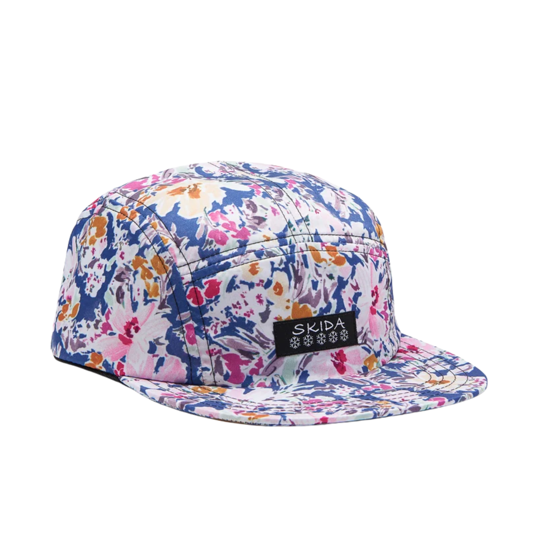 A five-panel camp cap with a vibrant floral print in blue, pink, orange, and white, featuring a black brand patch on the side with the text 'SKIDA' and snowflake logo.