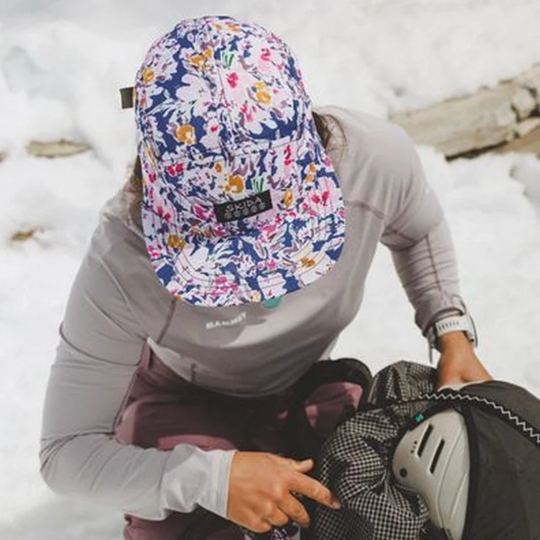 A person in a gray long-sleeve top and dark pants kneels in snow while wearing a floral-patterned baseball cap with a black brand patch that reads 'Skida'.