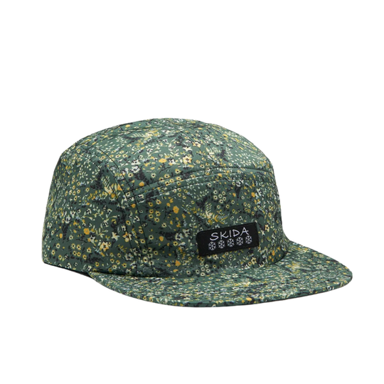 A five-panel camp cap in a dark green floral print with yellow and white flowers, featuring a black rectangular patch on the front with the white SKIDA logo.