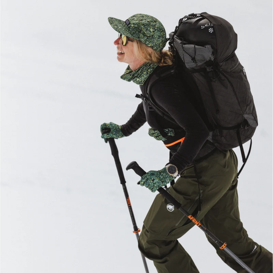 A woman hiking in the snow wearing a green camouflage hat, matching neck gaiter, gloves, and pants, along with a large black backpack and trekking poles.