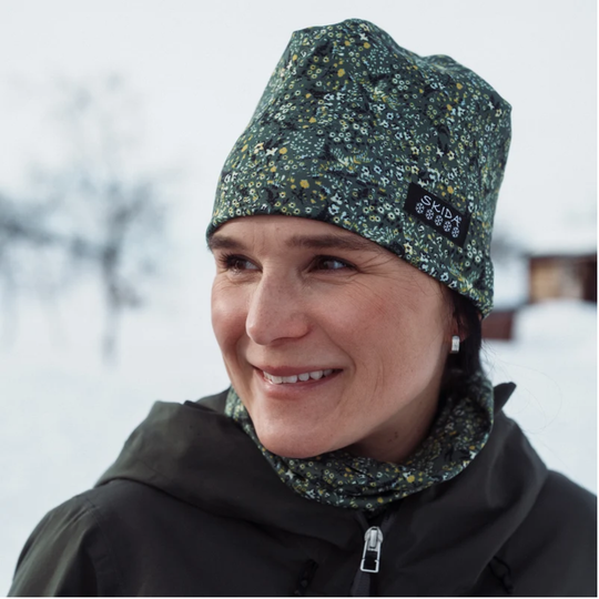 A woman wearing a dark green floral-patterned beanie and matching neck gaiter in a snowy outdoor setting. A black brand patch on the beanie reads 'skida'.