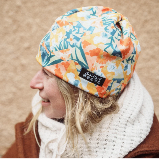 A woman wearing a colorful floral beanie with a black 'skida' brand tag, paired with a white knit scarf and brown jacket, set against a neutral beige background.
