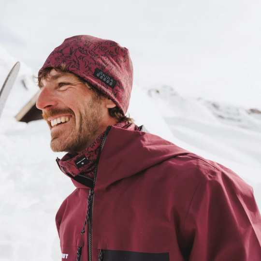 A man smiling outdoors in the snow, wearing a burgundy patterned beanie and a matching burgundy ski jacket with black accents.