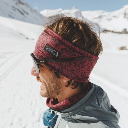 A man in a snowy mountain setting wearing a red patterned Skada headband and matching neck gaiter, along with black sunglasses.