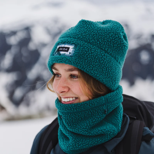 Skida High Pile Polartec Fleece Neckwarmer - Glade on a female model. Wearing a matching skida hat with snow and mountains in the background.