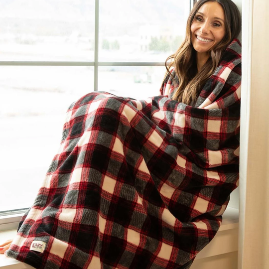 A woman wrapped in a red, black, and cream plaid blanket sits by a bright window. A small square brand patch reading 'lazy one' is visible near the lower left corner of the blanket.