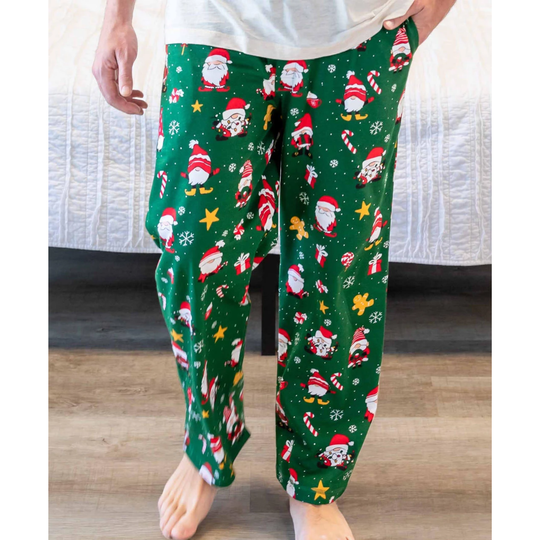 A person wearing green Christmas-themed lounge pants with a Santa Claus print, standing barefoot on a wood floor in front of a bed.