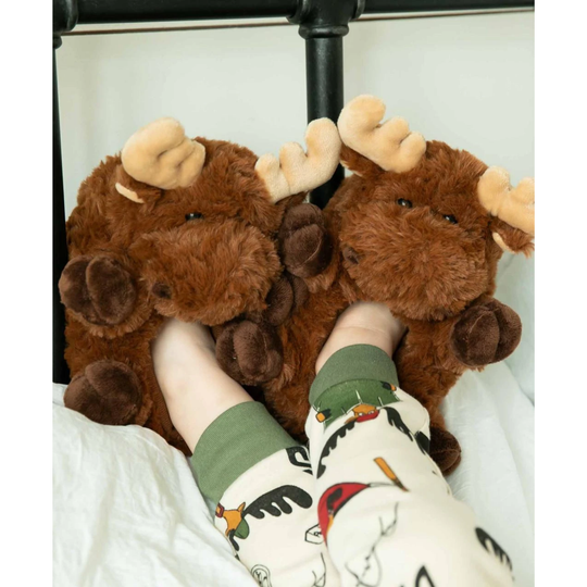 A person lying in bed with their feet tucked under two plush moose slippers. The slippers are brown with beige antlers and dark brown hooves, set against white bedding and a black bed frame.