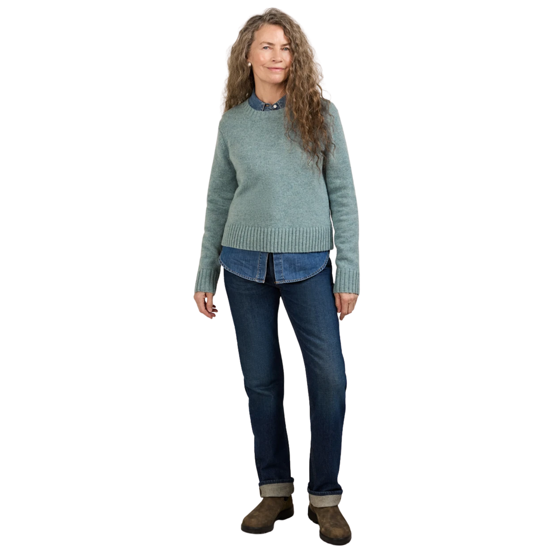 A woman modeling a light sage green knit sweater layered over a blue collared shirt, paired with dark blue jeans and brown boots, against a plain white background.