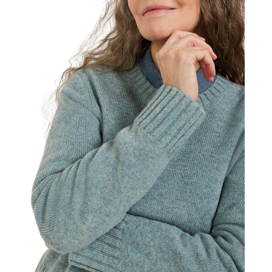 A close-up of a woman wearing a ribbed knit sweater in a dusty blue-gray color, with her hand resting near her chin against a plain white background.