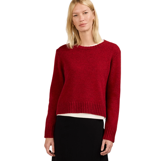 A blonde woman modeling a red knit crewneck sweater with long sleeves and ribbed cuffs, paired with a black skirt, against a plain white background.