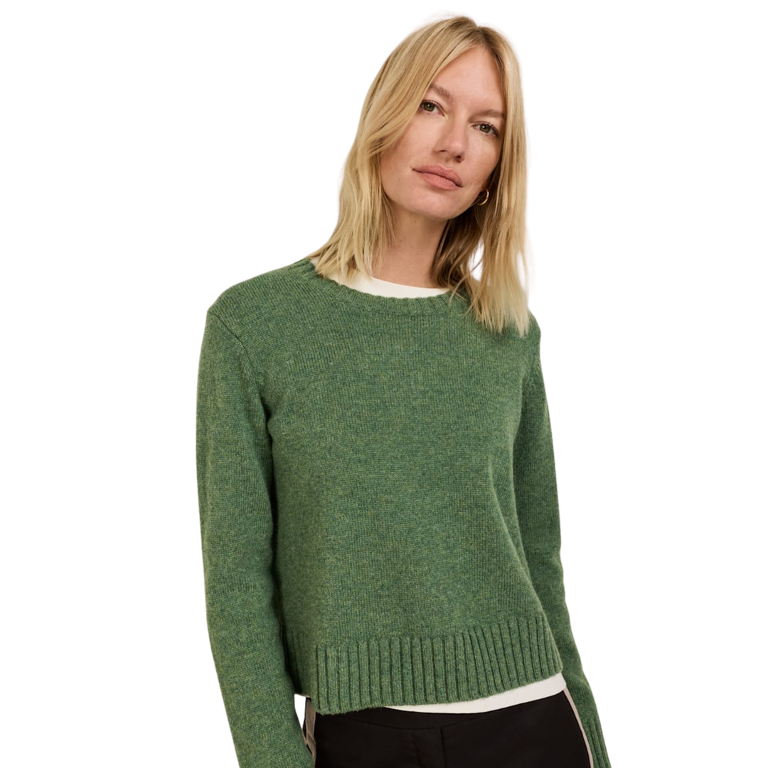 A blonde woman wearing a green knit crewneck sweater over a white top and black pants, photographed against a plain white studio background.