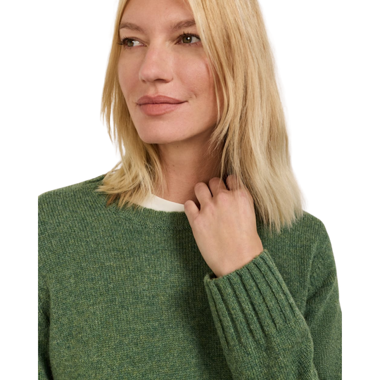 A blonde woman wearing a textured green knit sweater over a white undershirt, shown in a close-up profile shot against a plain white background.