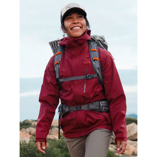 A woman hiking outdoors wearing a Marmot red jacket, gray backpack, and a beige cap, set against a natural rocky background.