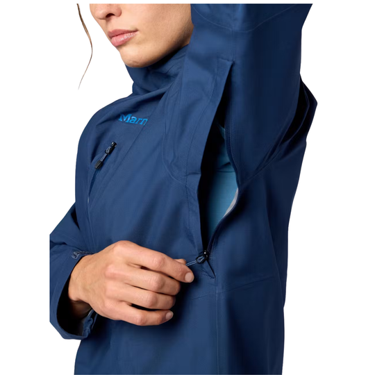 A close-up of a person wearing a navy blue technical outdoor jacket, showing the high collar, a zippered chest pocket with a blue 'Marmot' logo, and a hand opening a zippered sleeve against a white background.