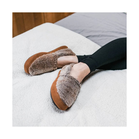 Brown fuzzy slip-on slippers worn with black leggings on a bed