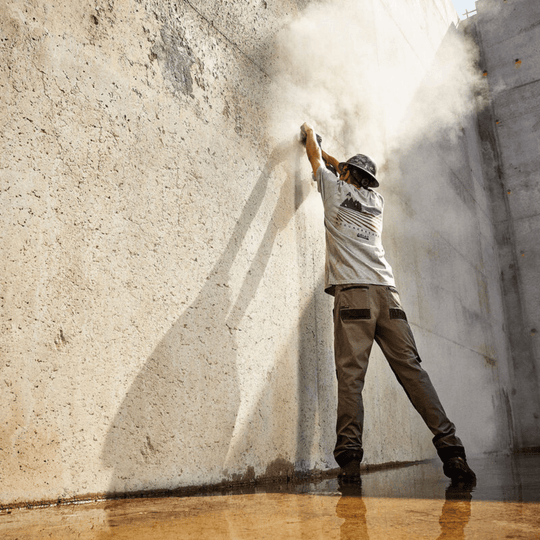 Tan straight-leg cargo pants with cargo pockets, worn by a person reaching up against a wall.