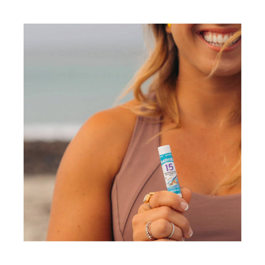 Lip balm tube labeled 15 with a blue-and-white cap, held by a smiling woman at the beach