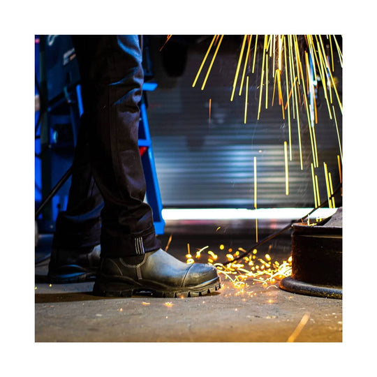 Black lace-up work boot with rugged treaded sole, shown in a workshop with sparks flying nearby