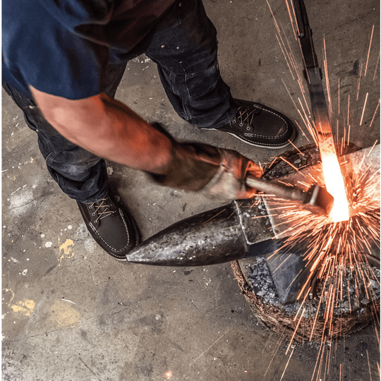 Dark moc-toe work boot with white stitching and laces shown beside an anvil and sparks