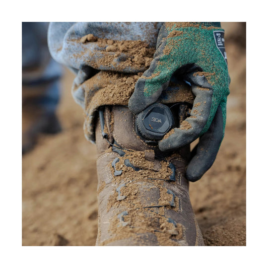 Mud-covered brown work boot with BOA dial being adjusted by a gloved hand