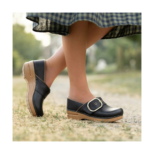 Black leather clog with wood-look sole and large buckle, shown in side view on a person walking outdoors