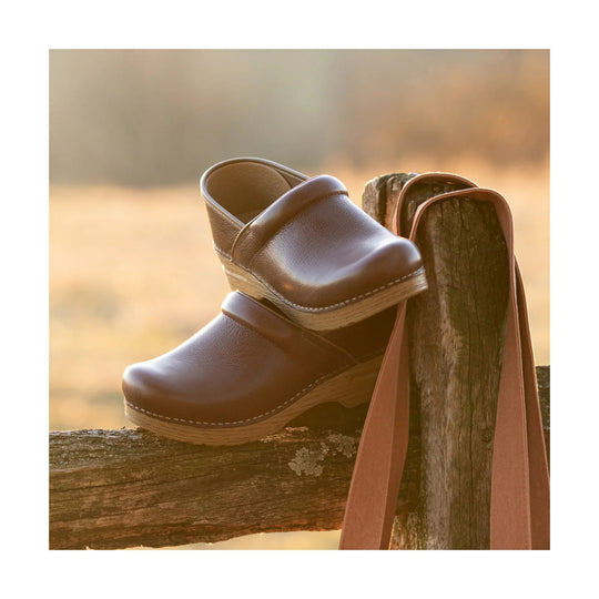 Brown clog-style slip-on shoes stacked on a wooden fence post with leather straps hanging beside them