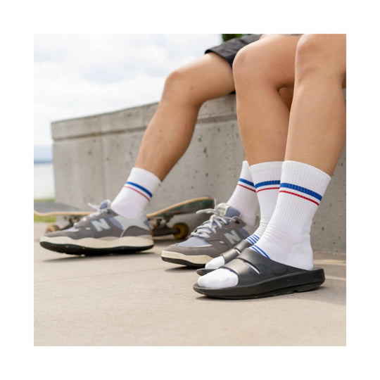 White crew socks with blue and red stripes worn with gray New Balance sneakers and black slide sandals