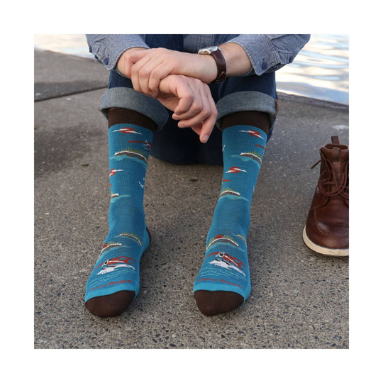 Blue crew socks with small airplane and landscape pattern, worn with rolled jeans and brown shoes nearby