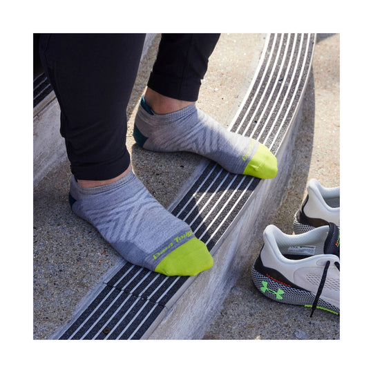 Gray no-show running socks with neon yellow toe and heel accents on a runner's feet, shown beside running shoes.