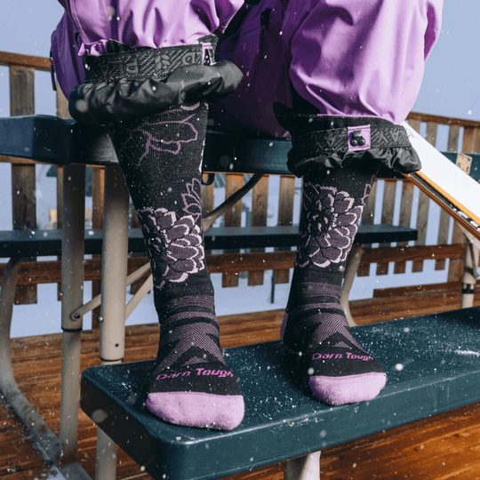 Black knee-high ski socks with pink floral pattern and pink toe text, shown on a person outdoors in snow