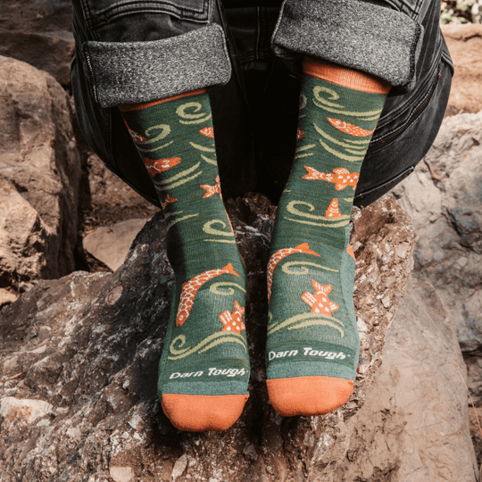 Green crew socks with orange fish pattern and "Darn Tough" text, worn with rolled jeans on rocks