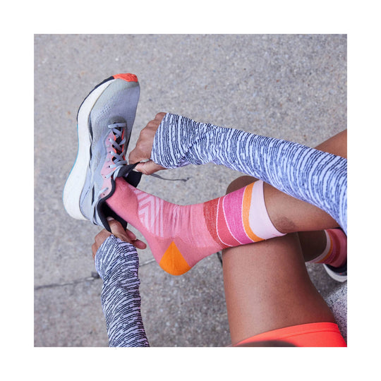 Light gray running shoe with pink accents and orange toe detail, being laced by a person wearing patterned arm sleeves and pink striped socks