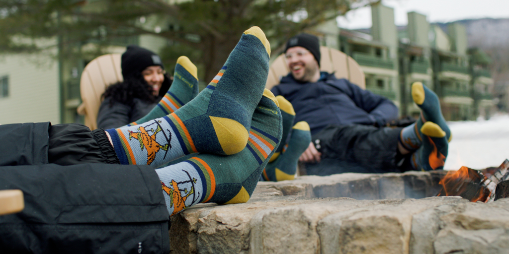 Two people sitting by a fire pit wearing colorful patterned socks with a scenic background.