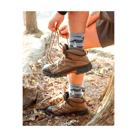 Tan hiking boots with black rubber toe caps being laced outdoors, worn with gray patterned crew socks.