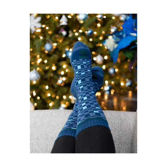 Blue patterned crew socks worn with black leggings, shown with crossed feet in front of a lit Christmas tree.
