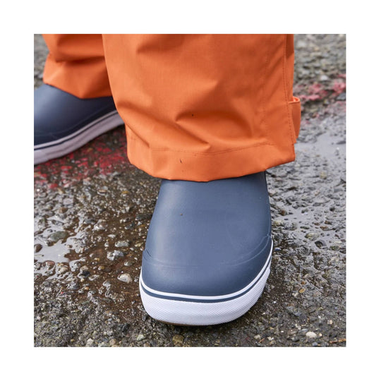 Navy slip-on rain boot with a white toe cap and textured sole, shown on wet pavement with orange pants