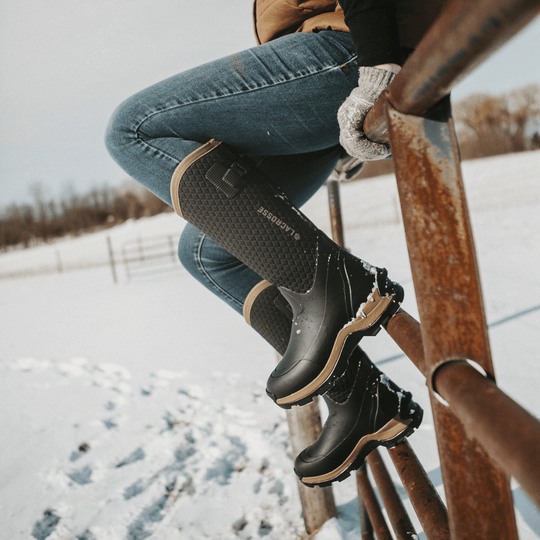 Black winter boot with tan trim and a textured side panel worn in snowy conditions