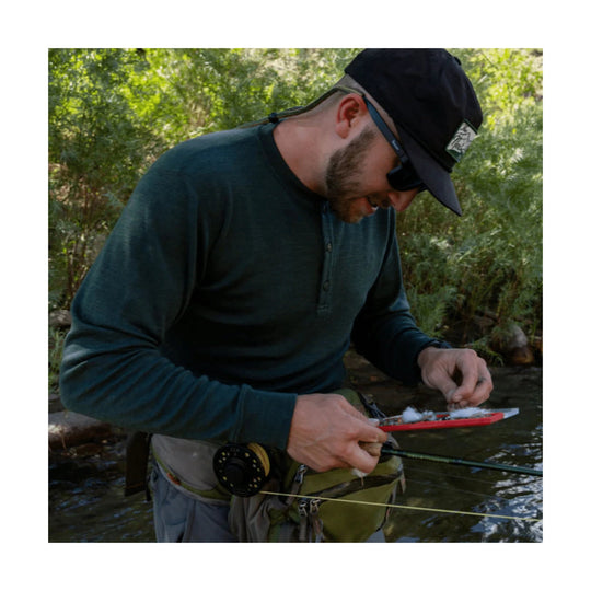 Man wearing a dark green henley shirt and black cap while fly fishing in a stream