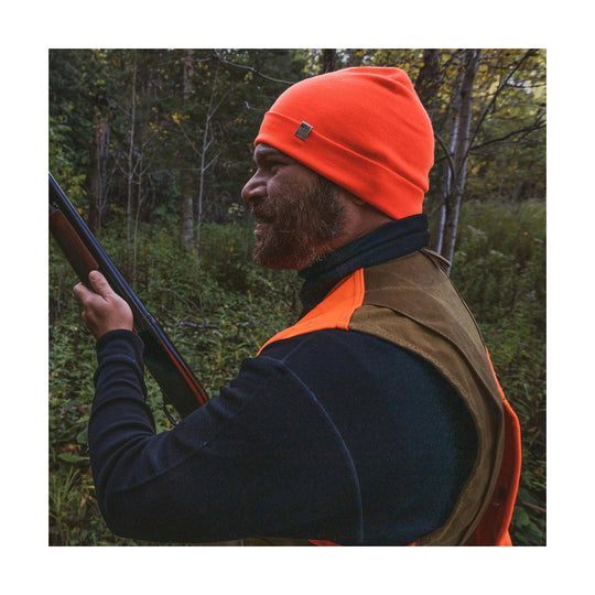 Man in orange beanie and dark turtleneck wearing a hunting vest, holding a shotgun in the woods