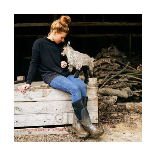 Tall mud boots on a seated woman petting a small goat