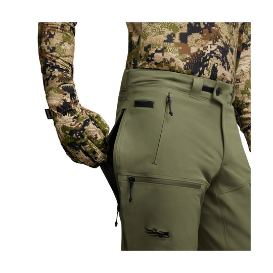 Olive green technical pants with zippered pockets and reinforced panels, shown on a model wearing camouflage gloves and top