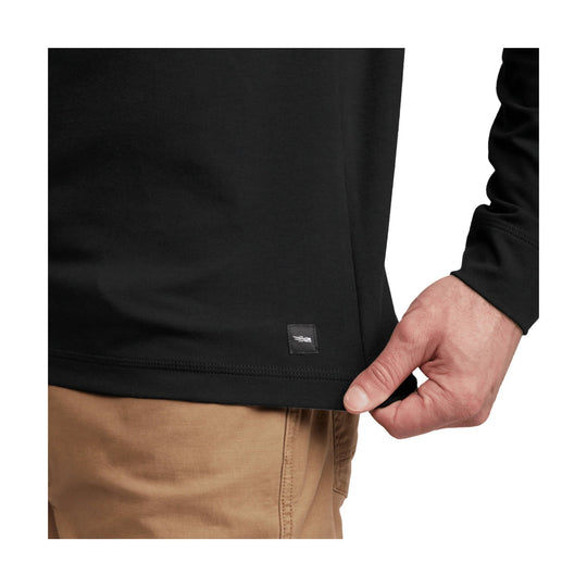 Black long-sleeve shirt hem with small woven tag, shown on a model with tan pants