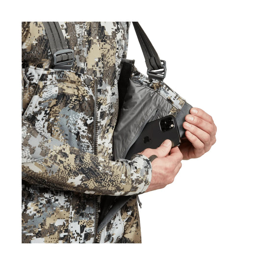 Camouflage hunting bib with front zipper pocket and shoulder straps, shown with a hand holding a phone inside
