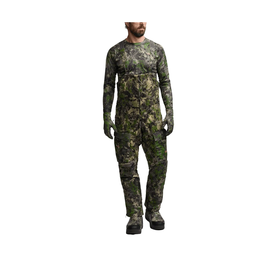 Camouflage hunting bib overalls with long sleeves, suspenders, cargo pockets, gloves, and boots