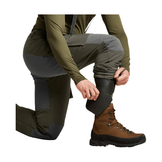 Crouching model wearing olive and gray hiking pants with reinforced knee panels and brown lace-up hiking boots