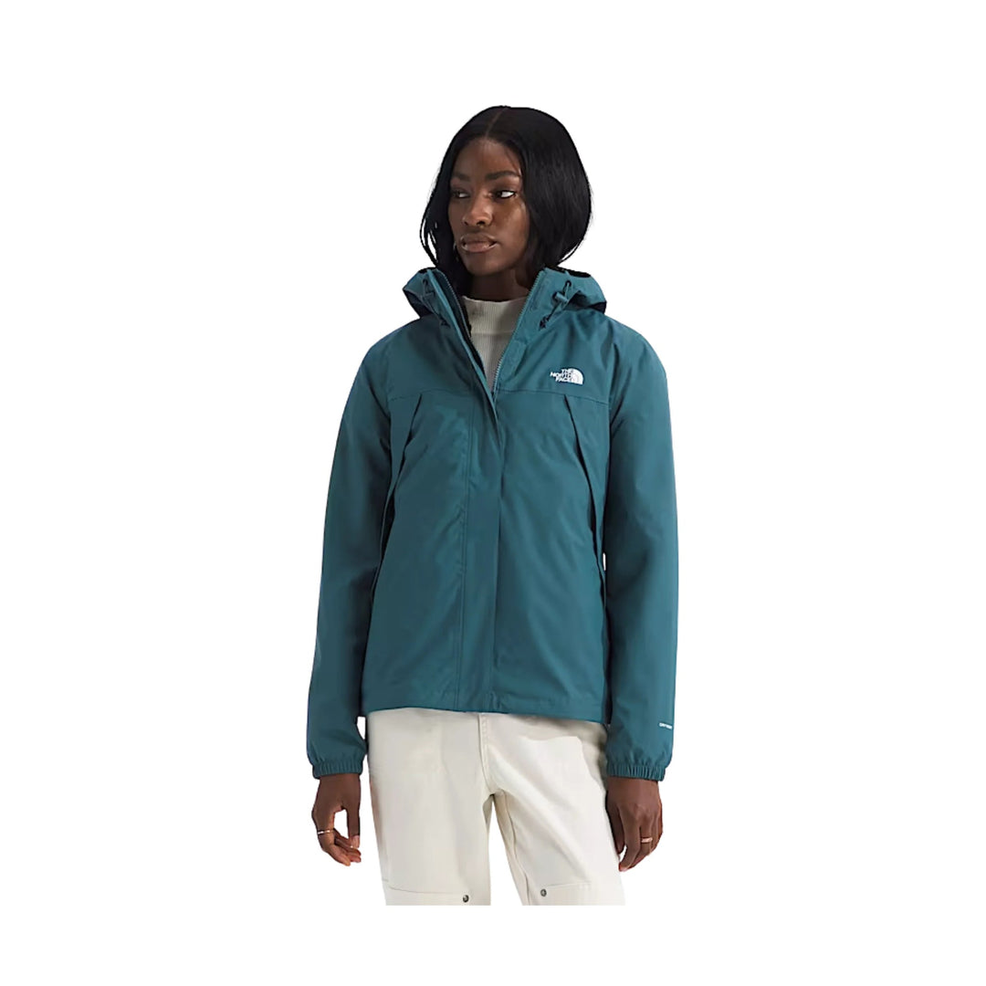 Teal hooded zip-up jacket with chest logo, worn over a light sweater and white pants