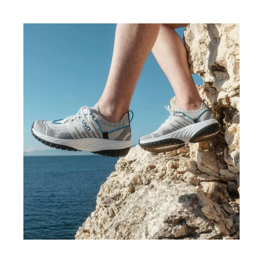 Light gray trail running shoes with blue accents and black tread, shown on a rocky seaside ledge