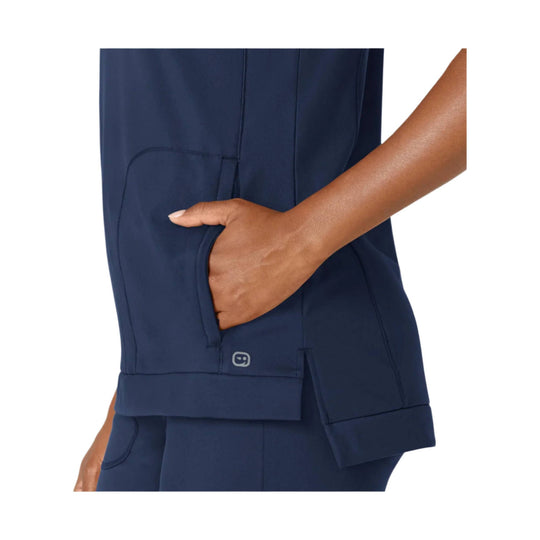 Navy scrub top with side pocket and small hem logo, shown on a model with hand in pocket