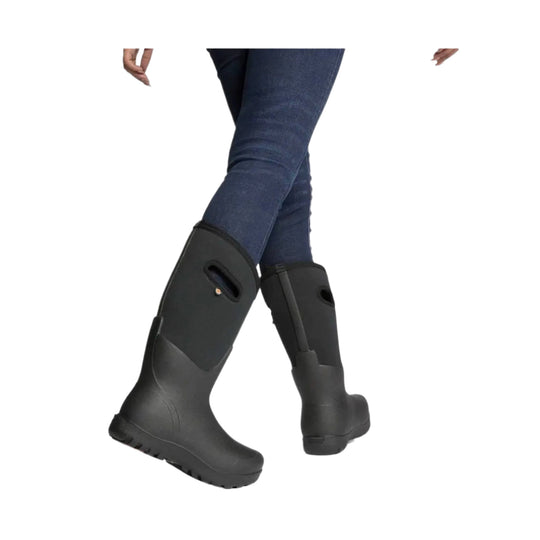 Black knee-high rain boots worn over dark blue jeans, shown in a crossed-leg view